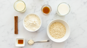 A flat lay of key ingredients for Cinnamon Roll Protein Crepes, including egg whites, protein powder, almond milk, oat flour, and cinnamon.